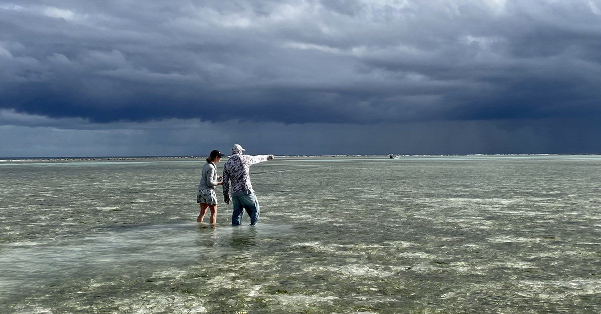 Guide pointing out a bonefish to a wading angler as a rain squall crosses the flat