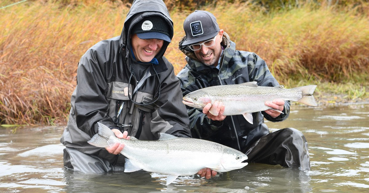 Two anglers kneeling in an Alaskan river holding a pair of rainbow trout, autumn grass behind them