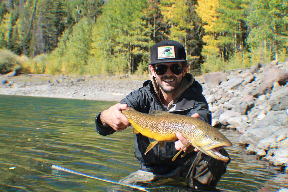 Angler holding a brown trout on a rocky shoreline with fall colors behind