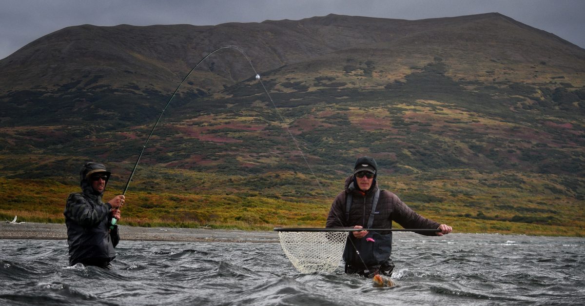 Two anglers wading a wind-ruffled river below a tundra mountain, one playing a fish