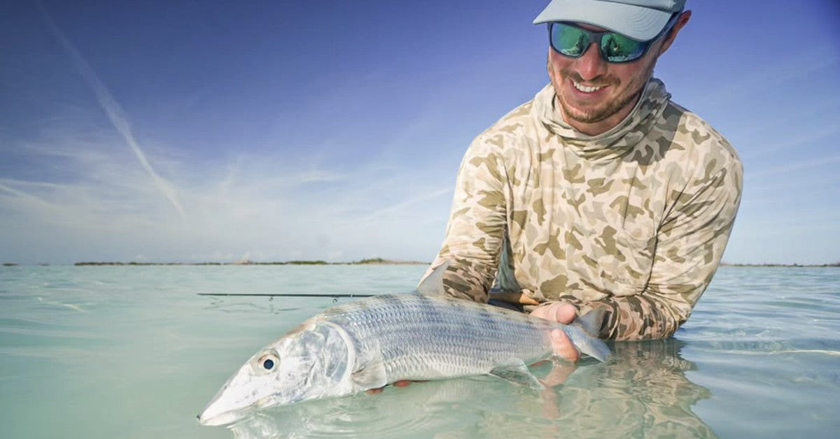 Angler in a camo sun hoody holding a bonefish just above the surface of a shallow tropical flat