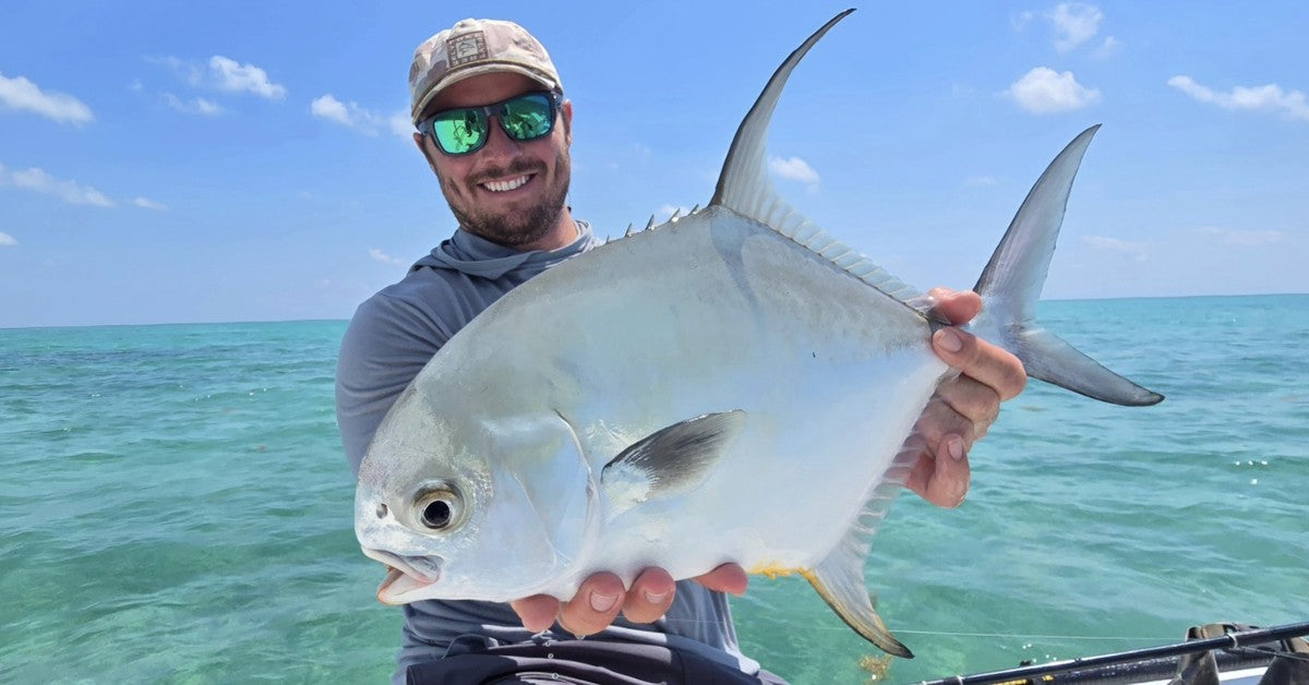 Angler on a skiff holding a permit horizontally over clear turquoise water, dorsal and tail fins fully visible