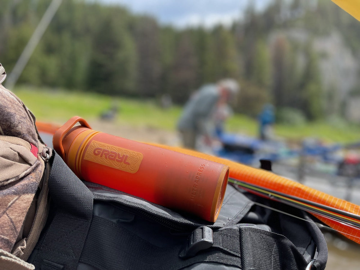Orange Grayl UltraPress purifier bottle resting on a pack beside a river with rods nearby