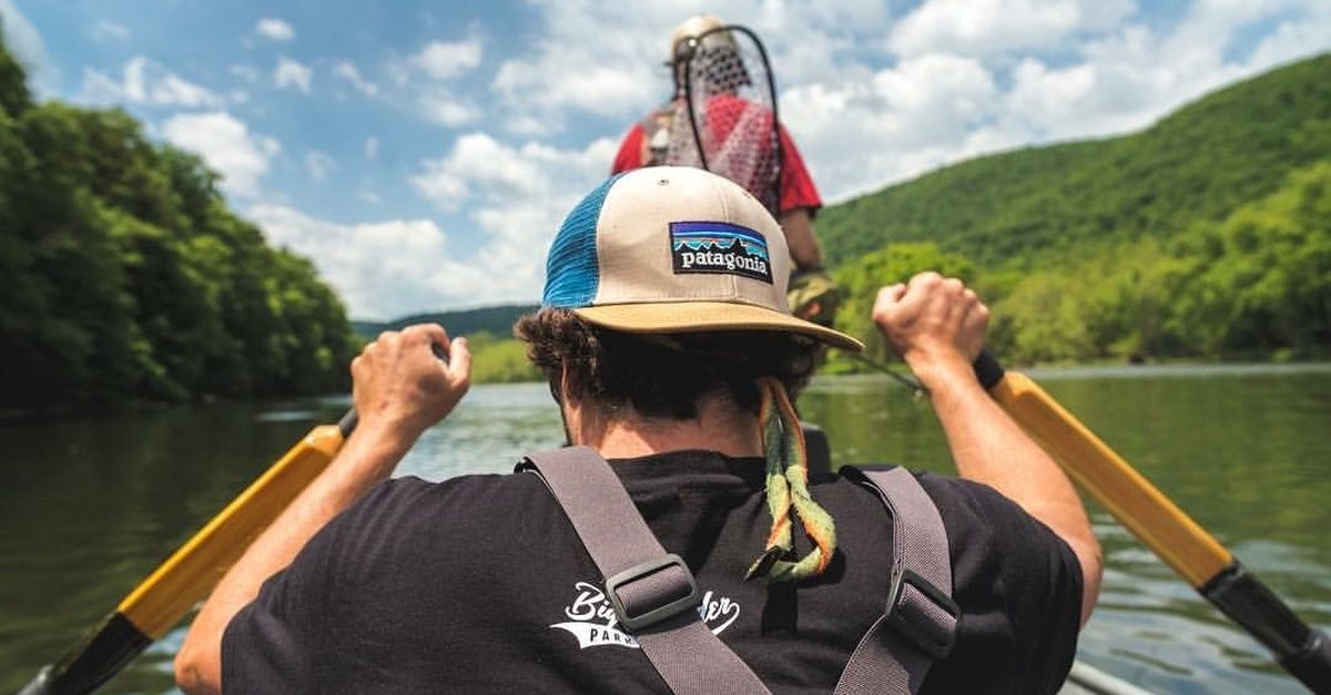 A guide pulling on the oars of a drift boat on a calm, tree-lined river
