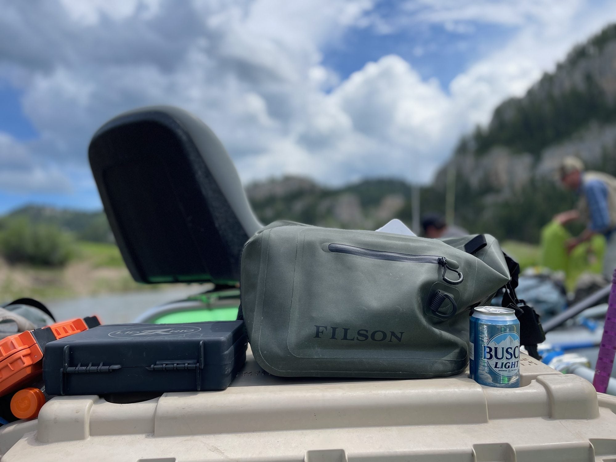 Olive Filson dry sling pack with a zippered front pocket sitting on a cooler lid next to a Lid Rig box on a raft