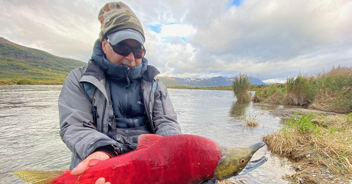 An angler kneeling on an Alaskan riverbank holding a sockeye salmon in full red spawning color, mountains behind