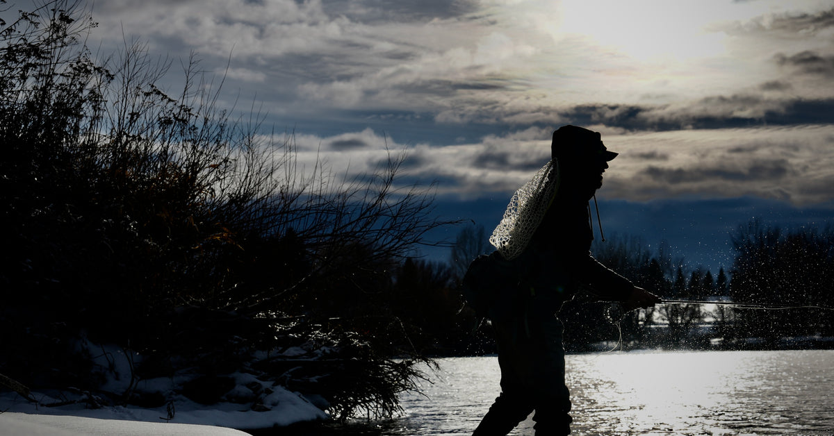 Angler wading through snow at dusk with a fly rod, silhouetted against fading light