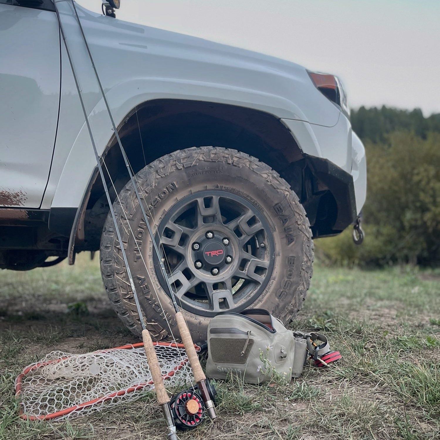 Simms hip pack, Rising Lunker net, Ross reel and Scott 4wt rod resting against a muddy truck tire at dusk