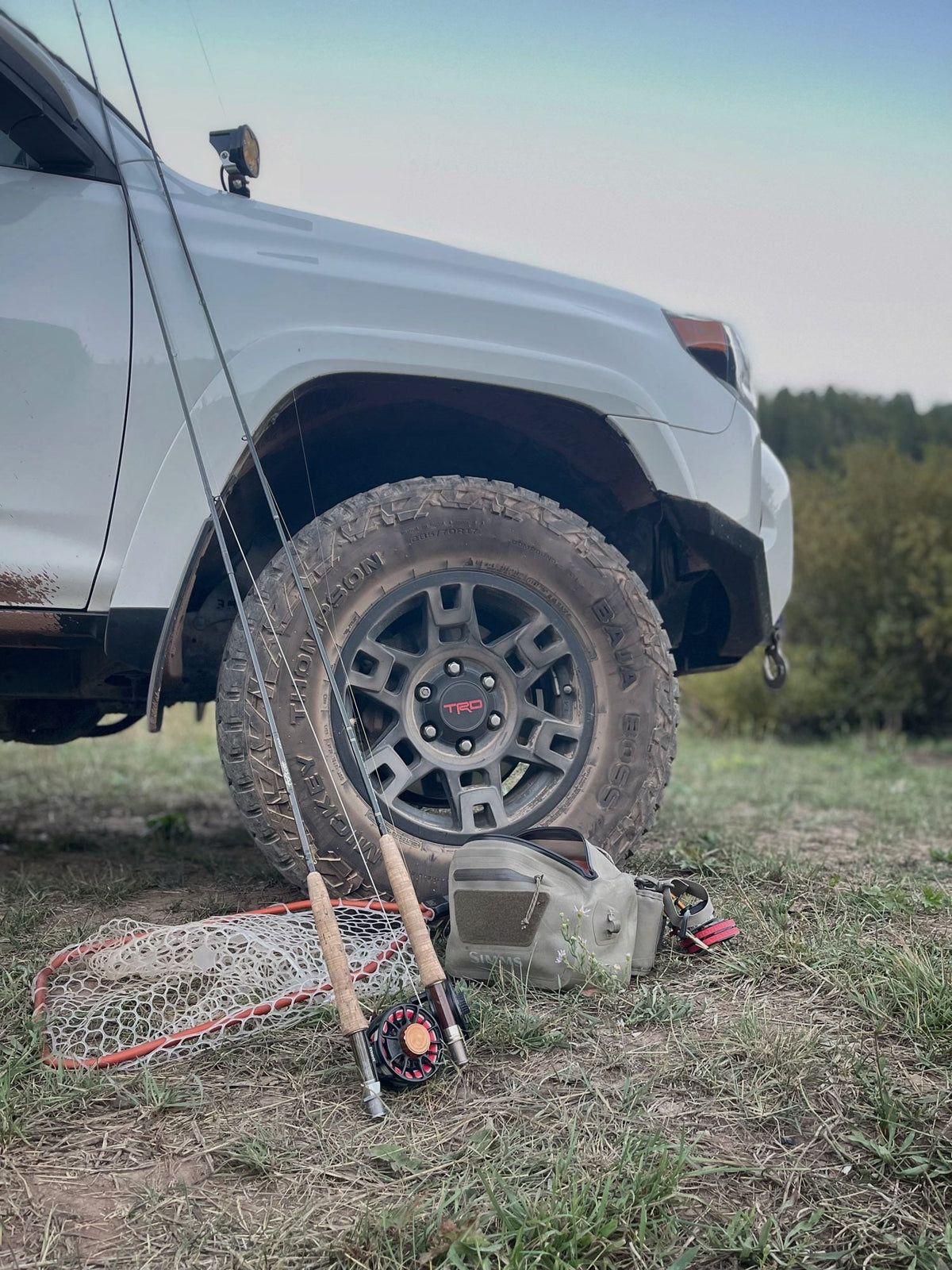 Simms hip pack, Rising Lunker net, Ross reel and Scott 4wt rod resting against a muddy truck tire at dusk