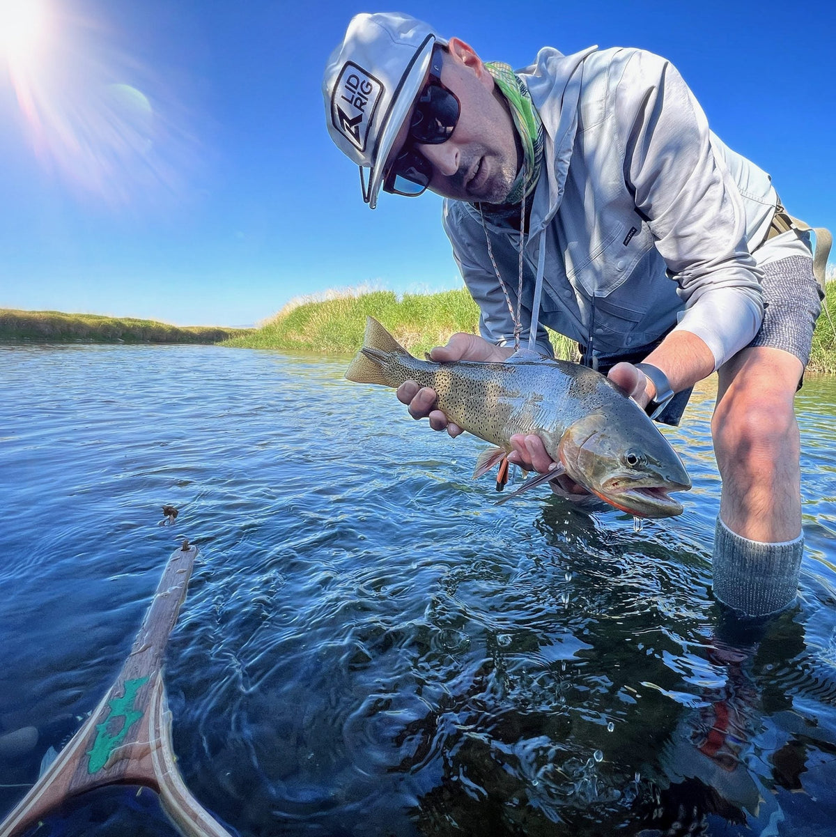 Angler holding a trout in a stream beside a wooden net with a green fish inlay on the handle