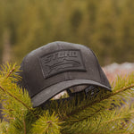 Dark gray trucker cap with STLHD logo patch, lying in pine needles