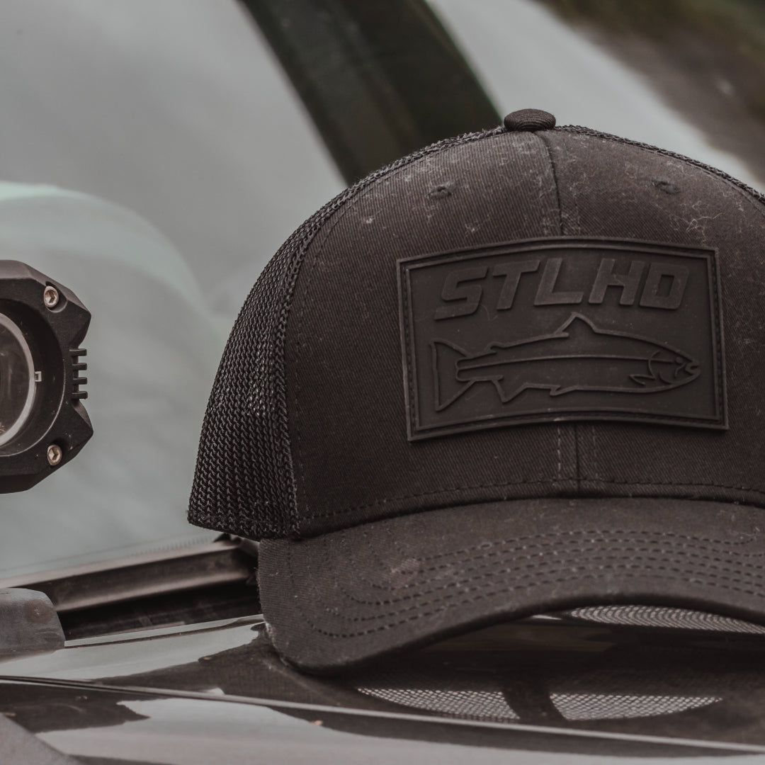 Dark trucker cap with STLHD logo patch on front panel, side view