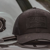 Dark trucker cap with STLHD logo patch on front panel, side view