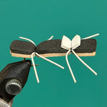 Ant-shaped decorative pin with black body, tan wings, and white leg ties